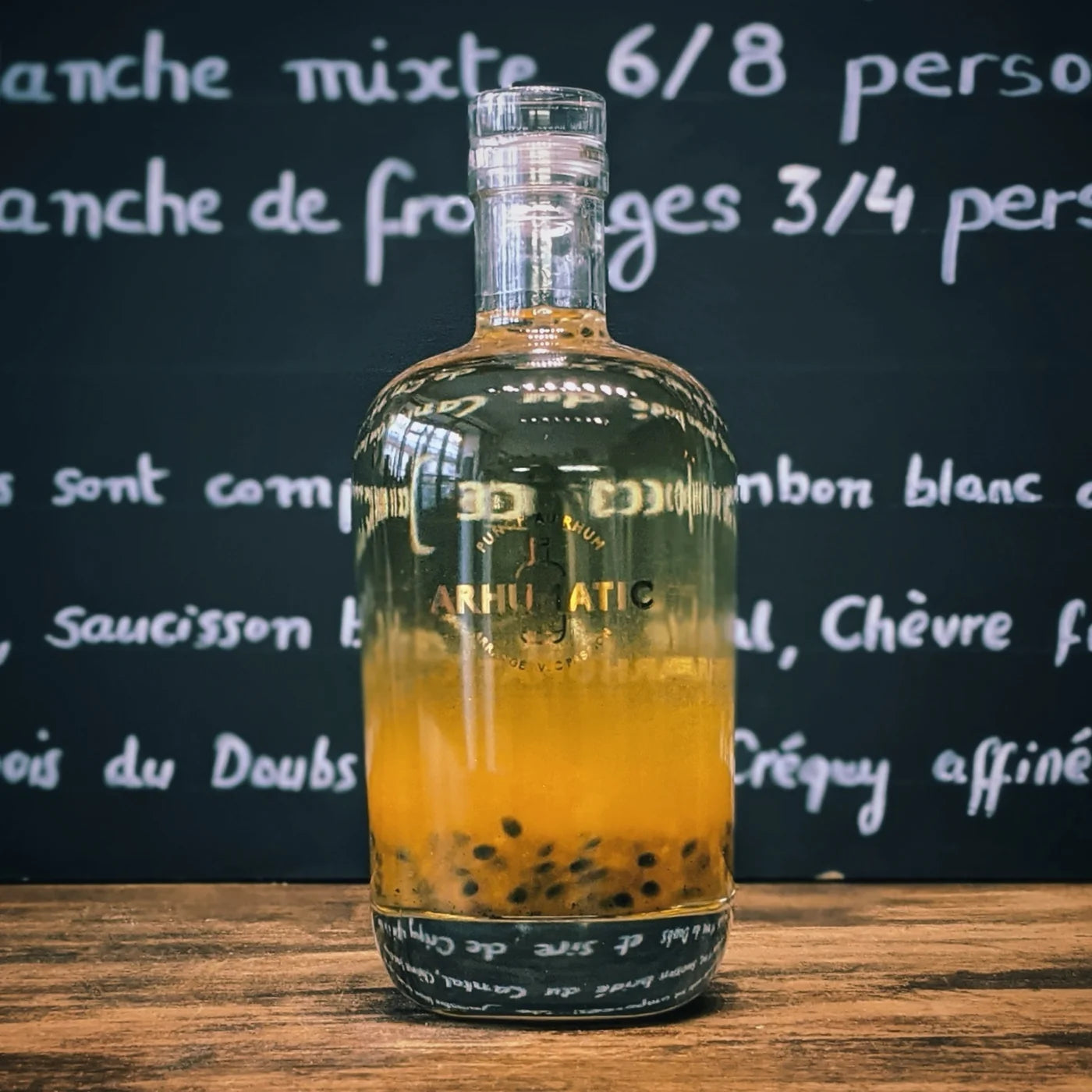 Bouteille d’Arhumatic Fruit de la Passion posée sur une table en bois, laissant apparaître la pulpe jaune et les graines noires du fruit dans le rhum, ambiance apéritif exotique et lumineuse.