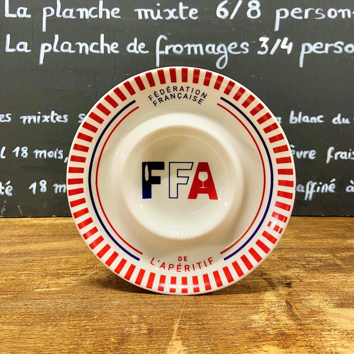 Assiette en porcelaine blanche FFA avec bord rouge et logo Fédération Française de l’Apéritif, posée sur une table en bois.