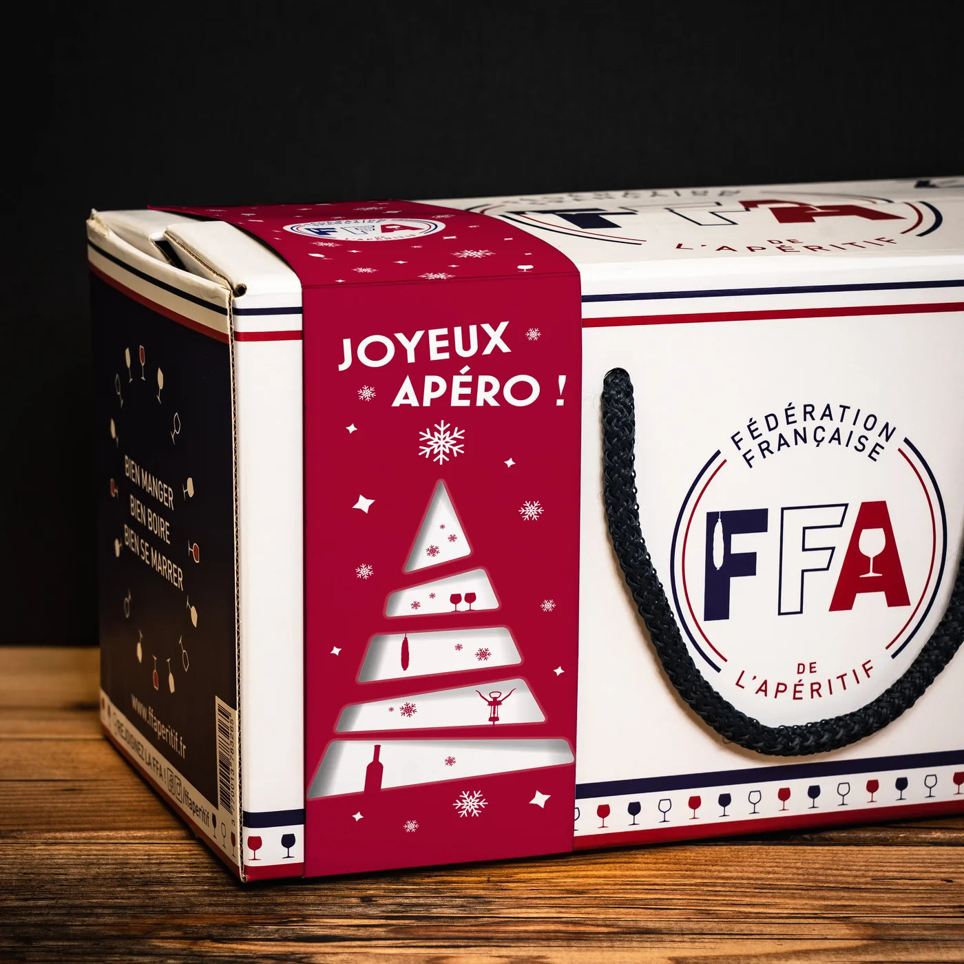 “Coffret L’Apér’Noël de la Fédération Française de l’Apéritif posé sur une table en bois, avec son design festif rouge et blanc — ambiance apéro conviviale et gourmande.”