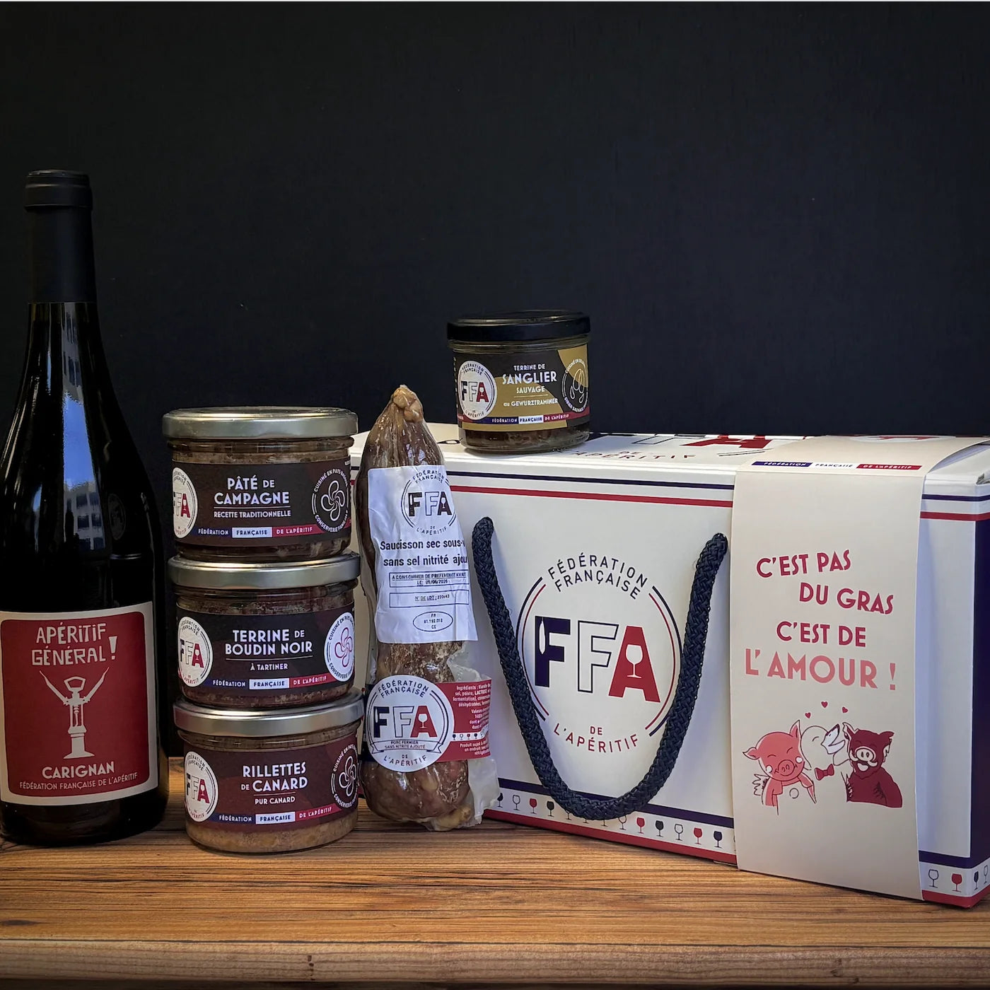 Coffret apéritif FFA avec vin rouge, terrines artisanales, rillettes de canard et saucisson sec, produits alignés devant un coffret cadeau au message humoristique.