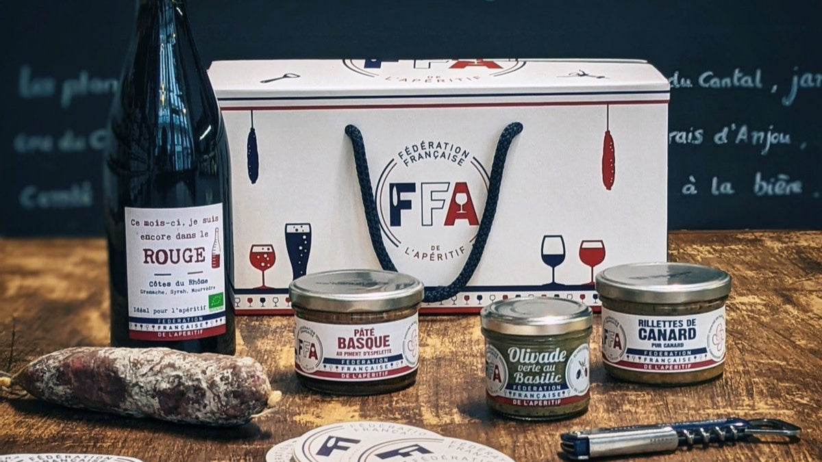 Coffret cadeau apéritif FFA avec vin rouge, rillettes, pâté basque, olivade verte et saucisson sur table en bois.