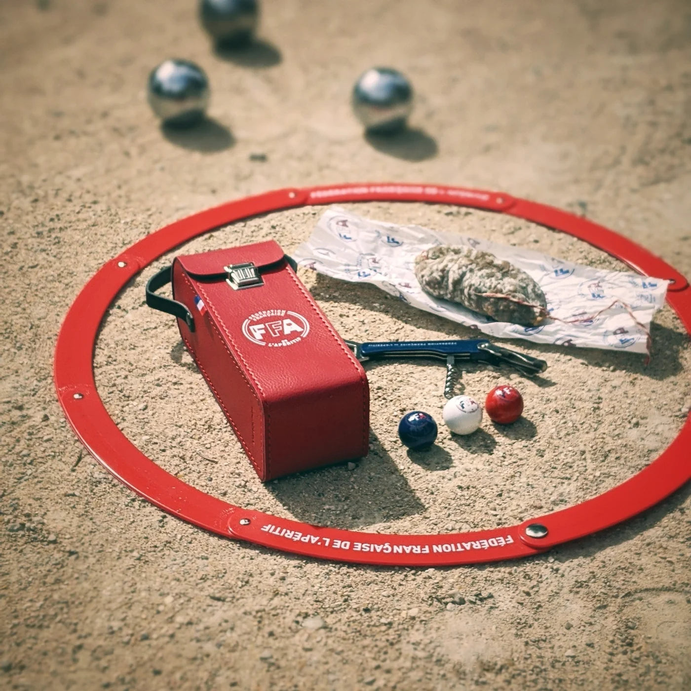 Étui rouge FFA pour 3 boules de pétanque posé dans un cercle de jeu sur le sable, avec un saucisson et des cochonnets tricolores — ambiance apéritif et partie de pétanque entre amis.