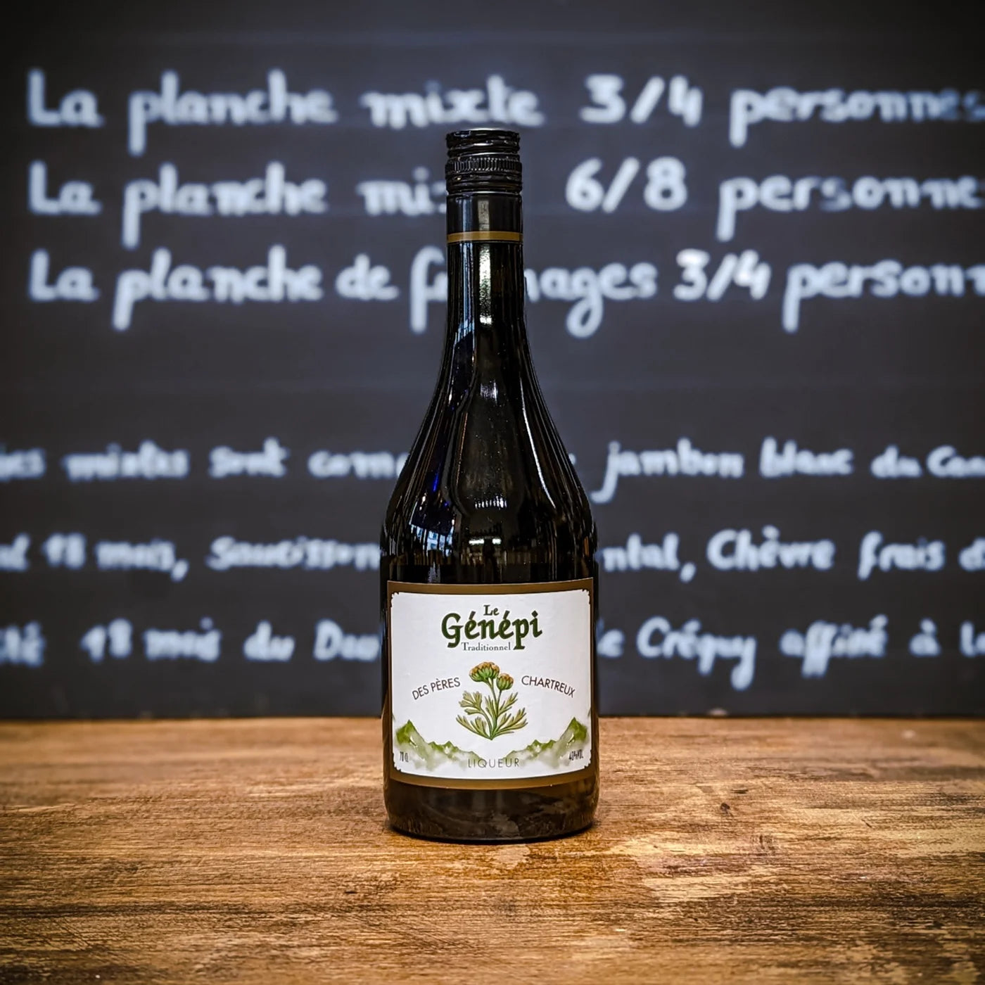Bouteille de liqueur de génépi des Pères Chartreux posée sur un comptoir en bois, avec tableau noir en arrière-plan, packshot apéritif artisanal.