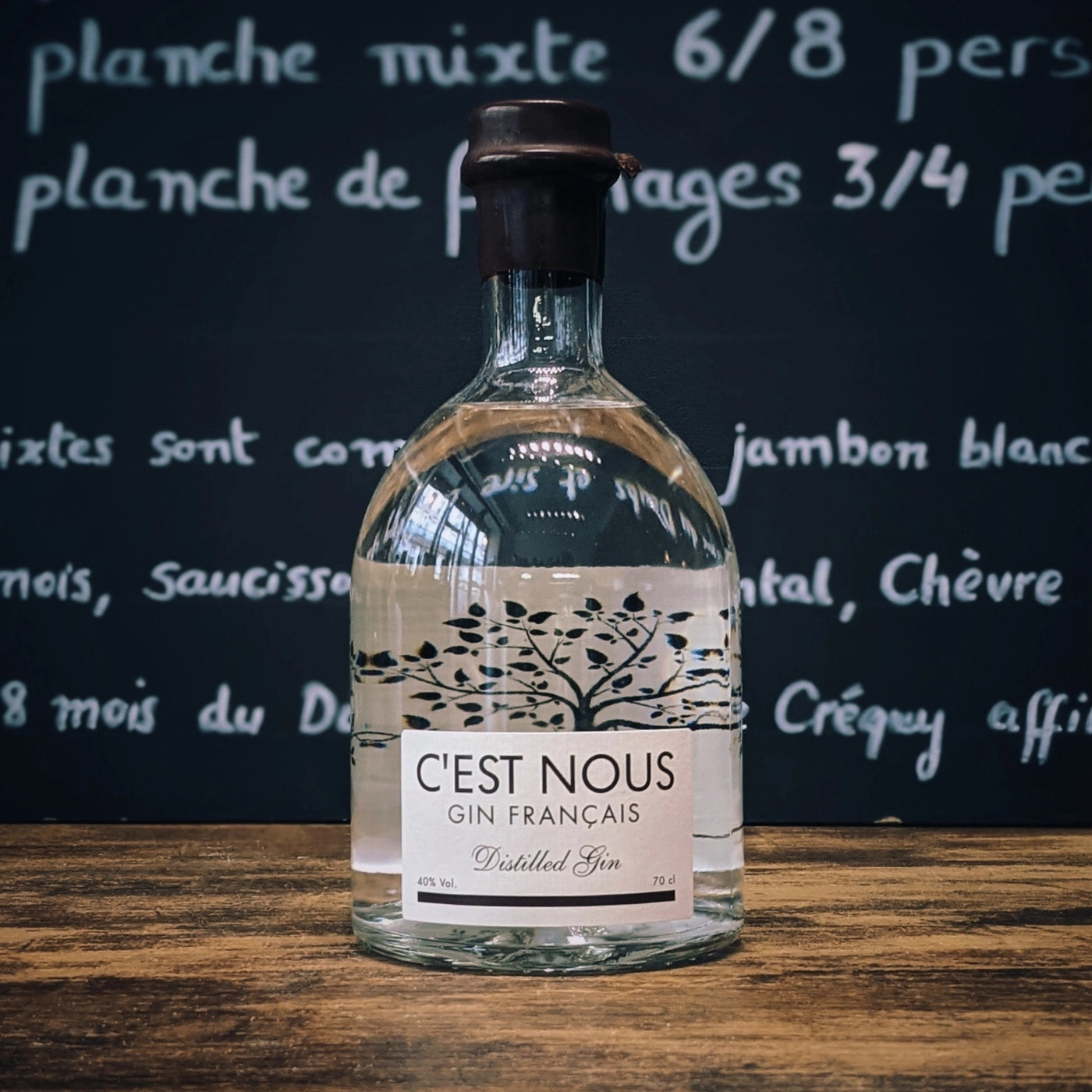 Bouteille de gin C’est Nous posée sur une table en bois avec un fond ardoise, ambiance apéritif française élégante et contemporaine.