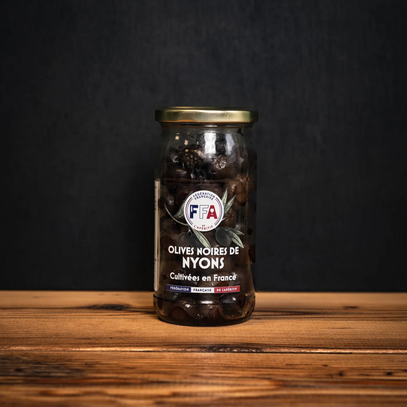 Bocal d’olives noires de Nyons FFA posé sur une table en bois, étiquette artisanale sur fond sombre — produit français gourmand pour l’apéritif.