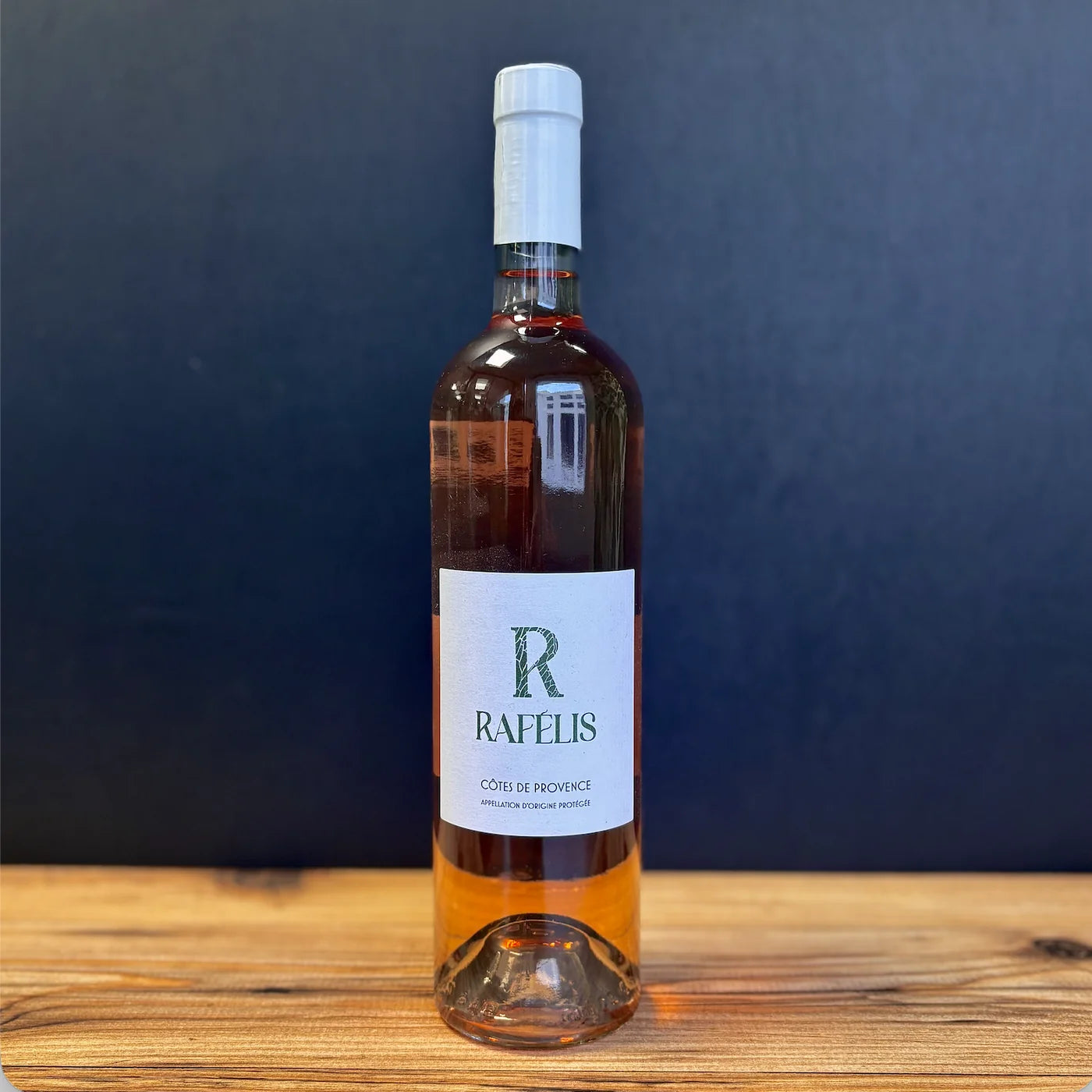 Bouteille de vin rosé Côtes de Provence Rapélis, posée sur une table en bois, fond bleu foncé, ambiance apéritive élégante et naturelle.