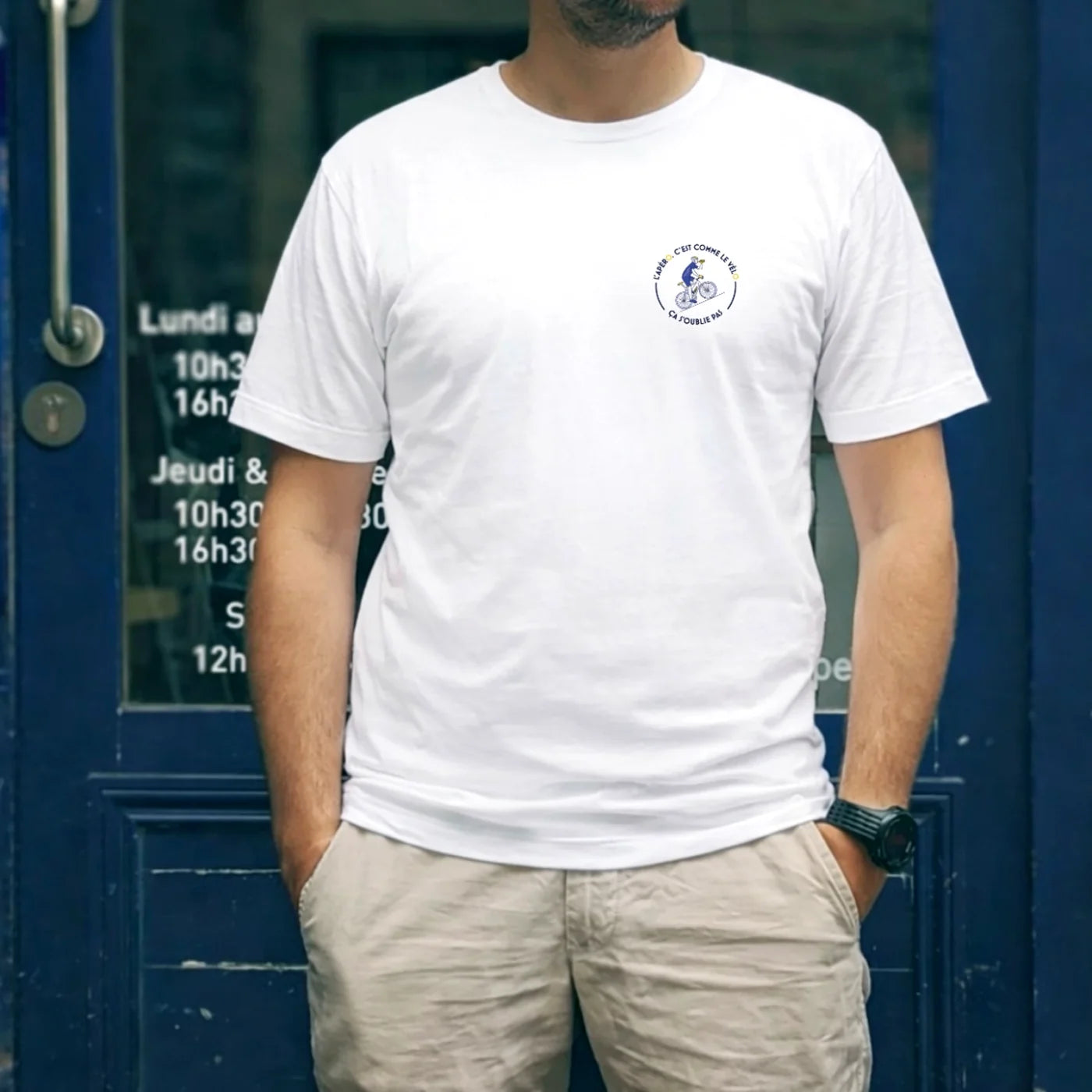 T-shirt blanc FFA “L’apéro c’est comme le vélo — ça s’oublie pas !” porté devant une devanture bleue, style apéritif cycliste.