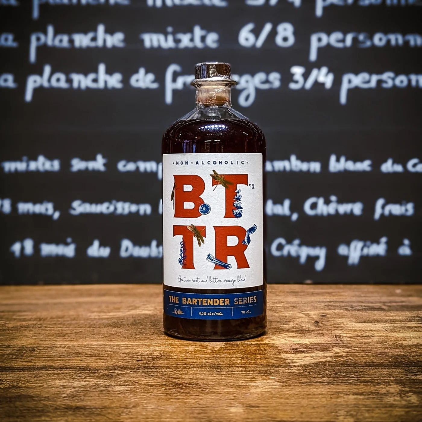 Bouteille BTR The Bartender Series posée sur un comptoir en bois, étiquette épurée bleu et rouge, ambiance de bar chaleureuse et gourmande pour un apéritif sans alcool premium.
