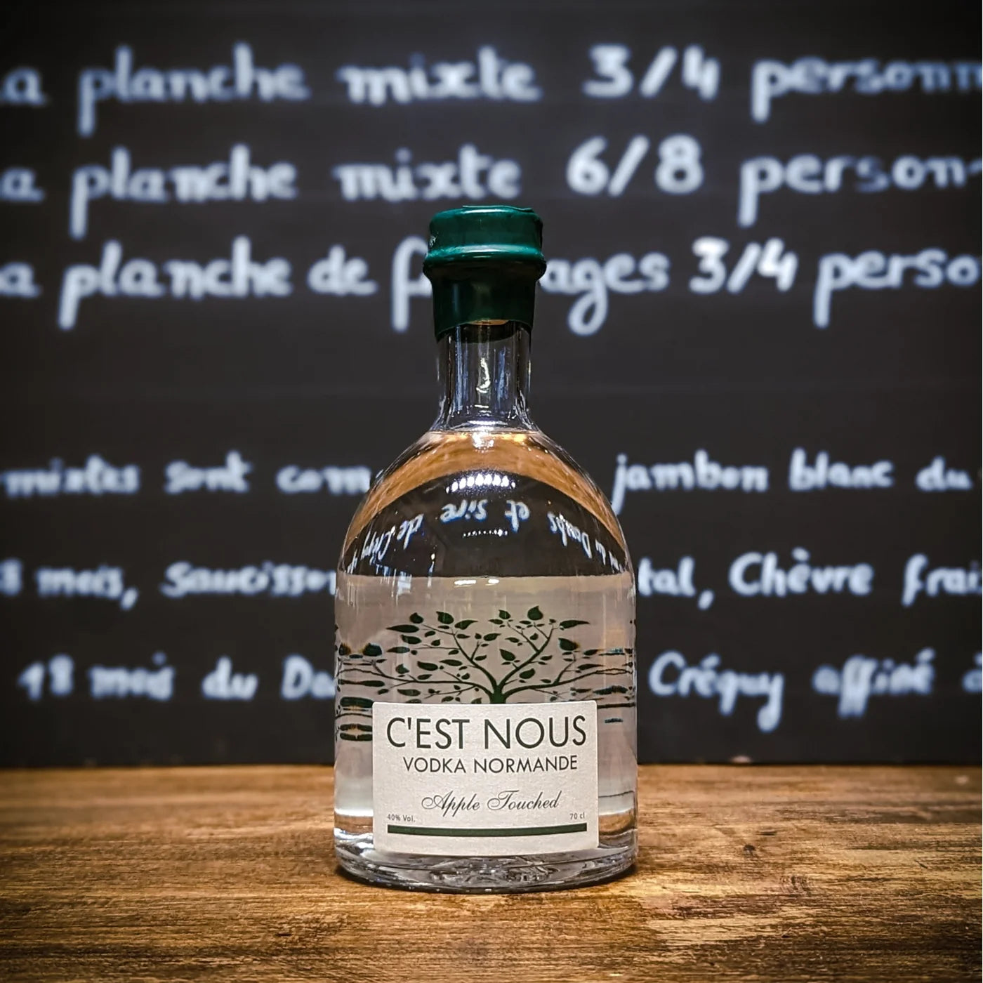 Bouteille de vodka normande C’est Nous photographiée sur un comptoir en bois devant un tableau noir, packshot apéritif premium.