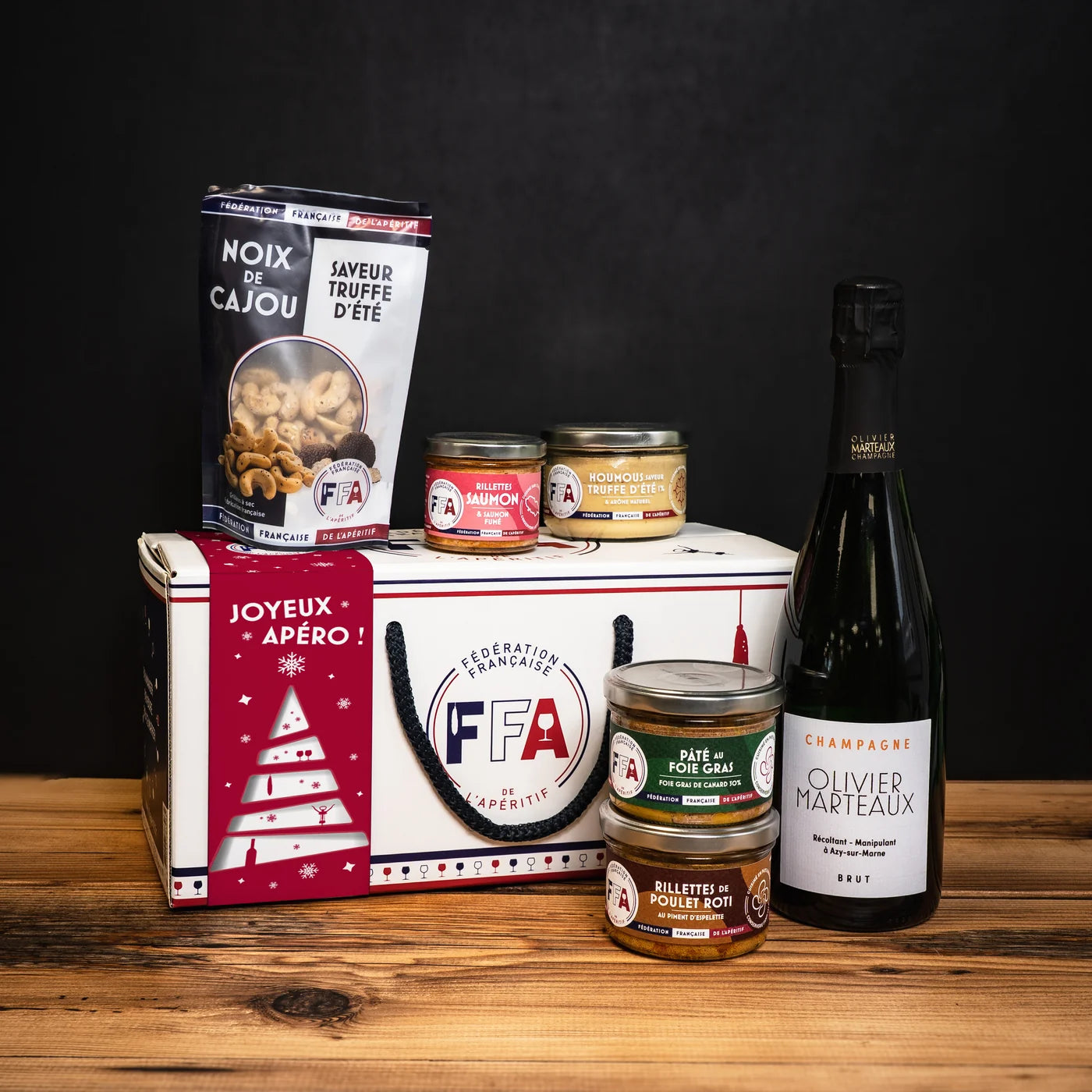 Coffret L’Apér’Noël FFA entouré de produits apéritifs artisanaux et d’une bouteille de champagne, mis en scène sur une table en bois dans une ambiance chaleureuse.