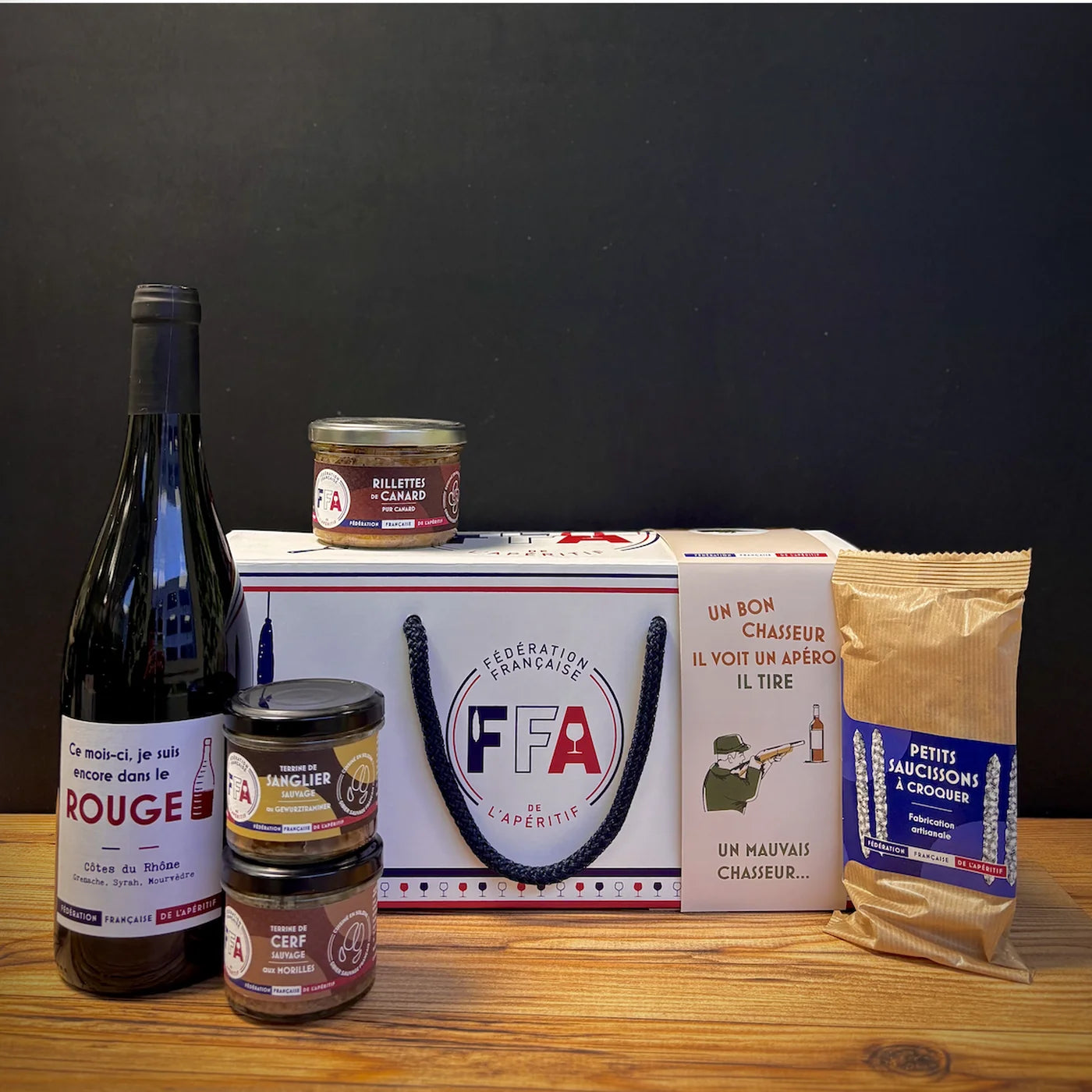 Coffret apéritif FFA thème chasse avec vin rouge, terrines de gibier, rillettes artisanales et saucissons à croquer, présenté sur une table en bois.