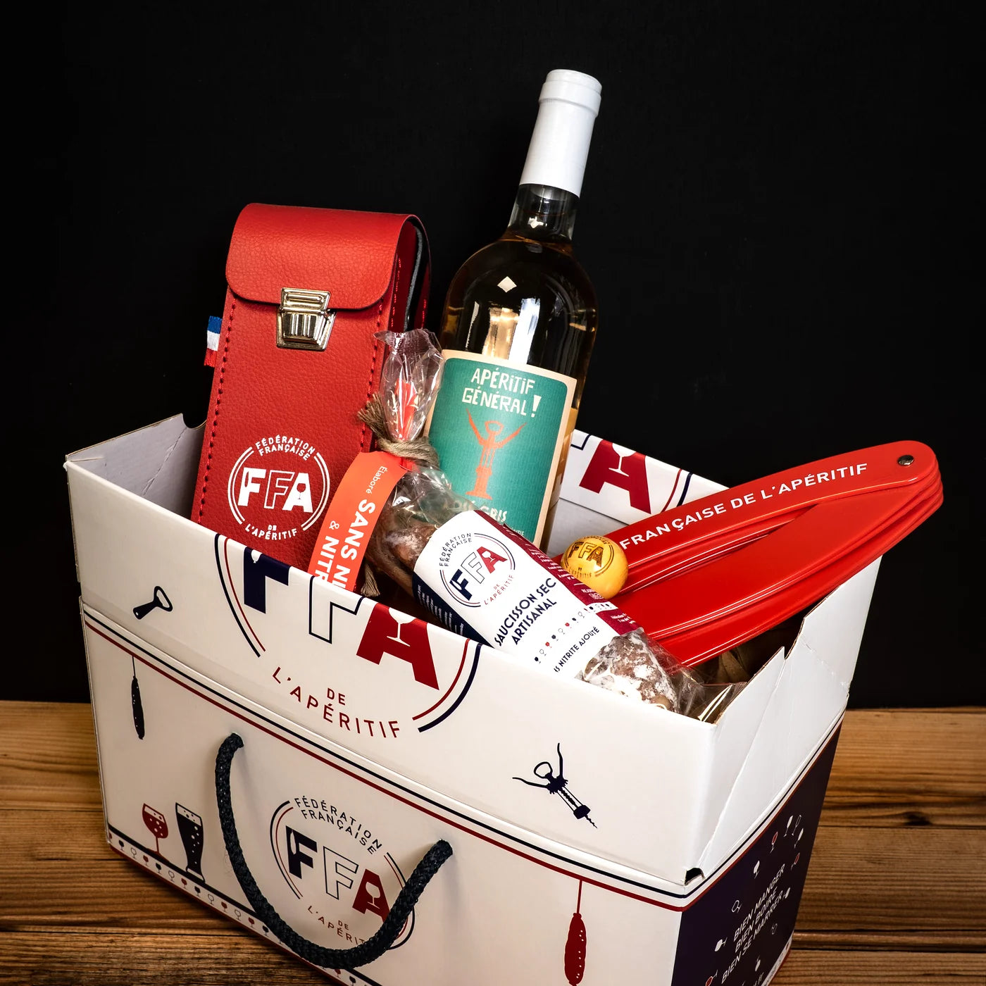Coffret apéritif FFA rempli de produits artisanaux — bouteille de rosé gris, saucisson, tapenade et accessoires, ambiance bois et fond sombre.