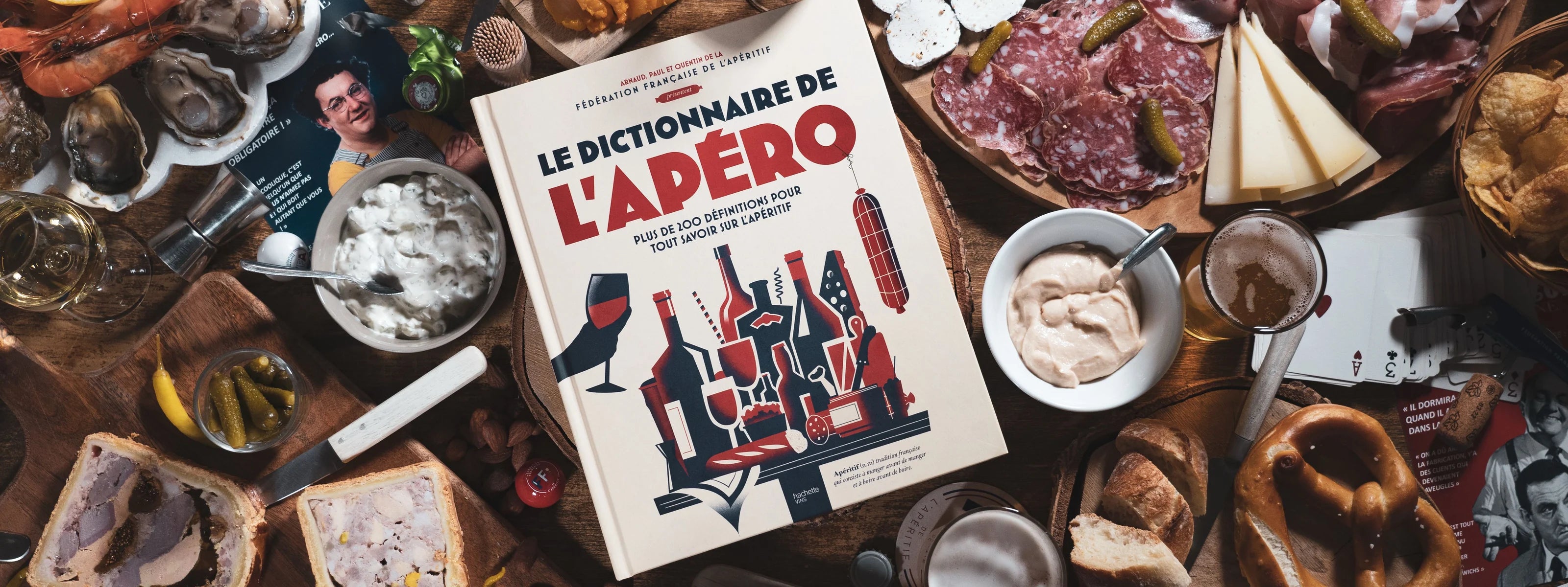 Le Dictionnaire de l’Apéro FFA posé au centre d’une grande table garnie de charcuterie, fromages, fruits de mer et vins — ambiance conviviale et gourmande d’apéritif français.