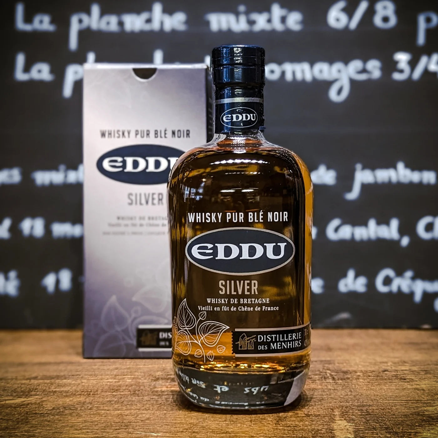 Bouteille de whisky Eddu Silver posée sur un comptoir en bois, avec son étui en arrière-plan — whisky breton pur blé noir, mise en scène épicerie fine.