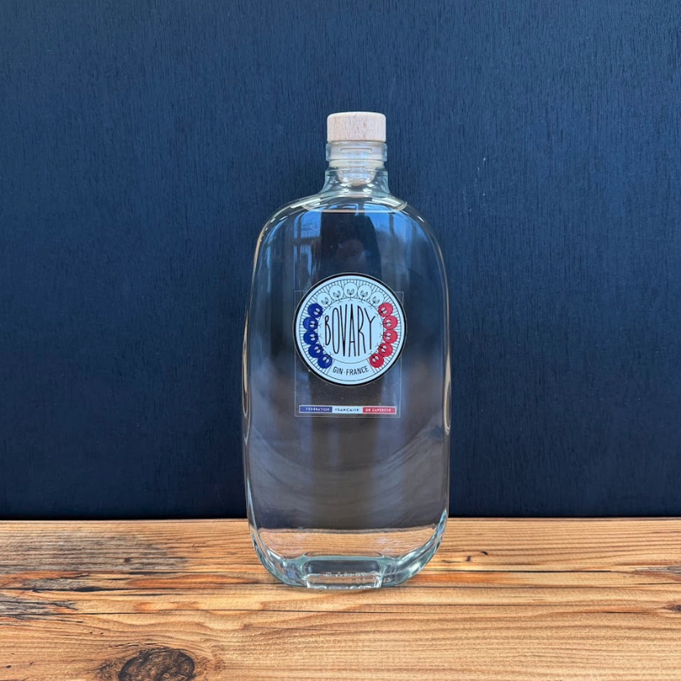 Bouteille de Gin Bovary de la Fédération Française de l’Apéritif posée sur un fond noir avec une table en bois clair, mise en scène lumineuse et épurée.