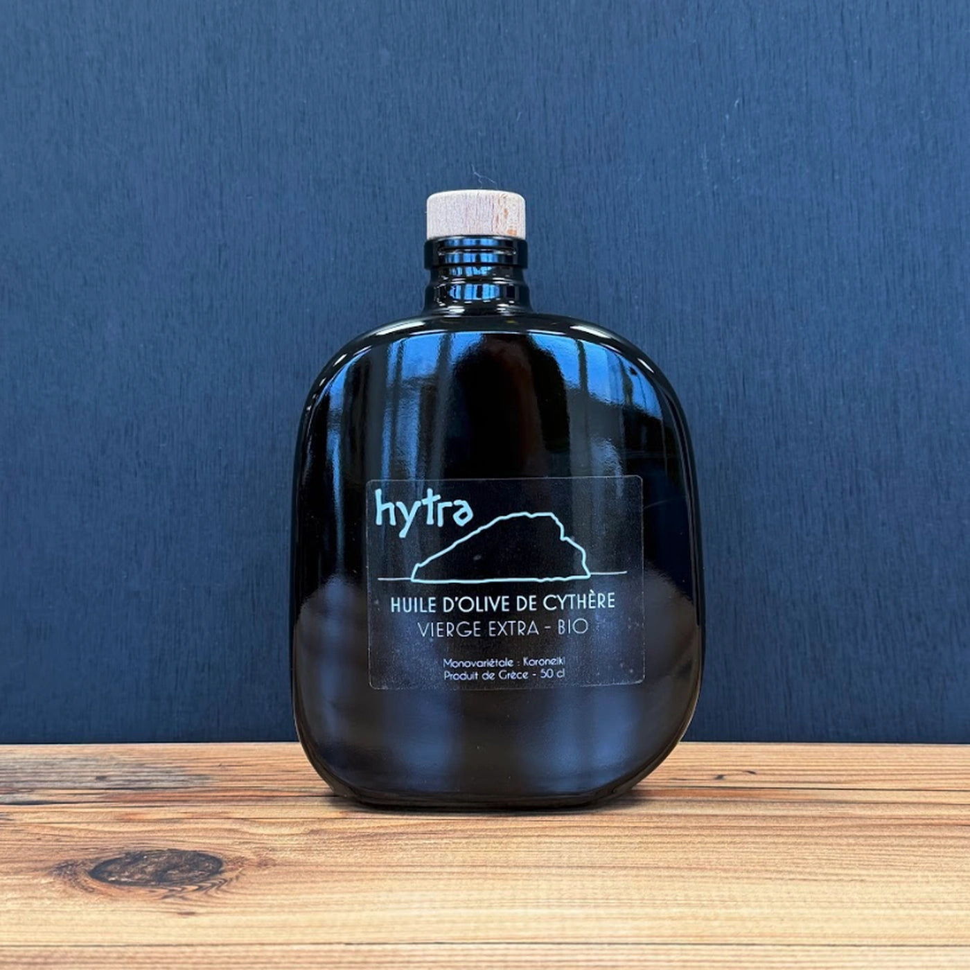 Bouteille d’huile d’olive Hytra posée sur une table en bois clair, fond noir élégant, reflet naturel sur le verre, ambiance premium et méditerranéenne.