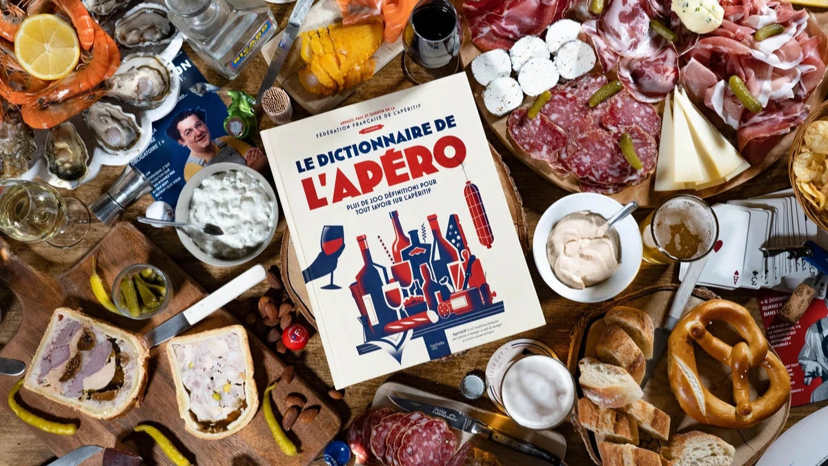 Le Dictionnaire de l’Apéro - livre officiel de la Fédération Française de l’Apéritif entouré de charcuteries, fromages et boissons. ￼