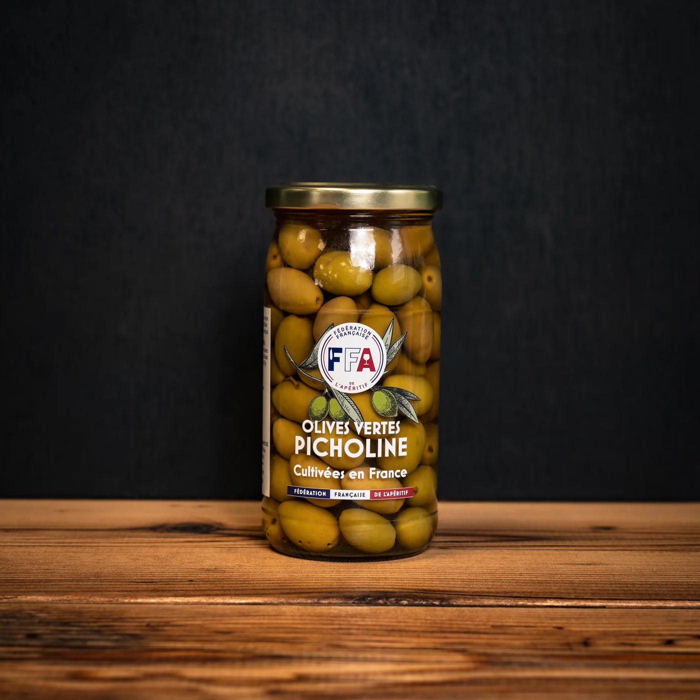 Bocal d’olives vertes Picholine FFA posé sur une table en bois, étiquette artisanale sur fond sombre — produit français authentique pour l’apéritif.