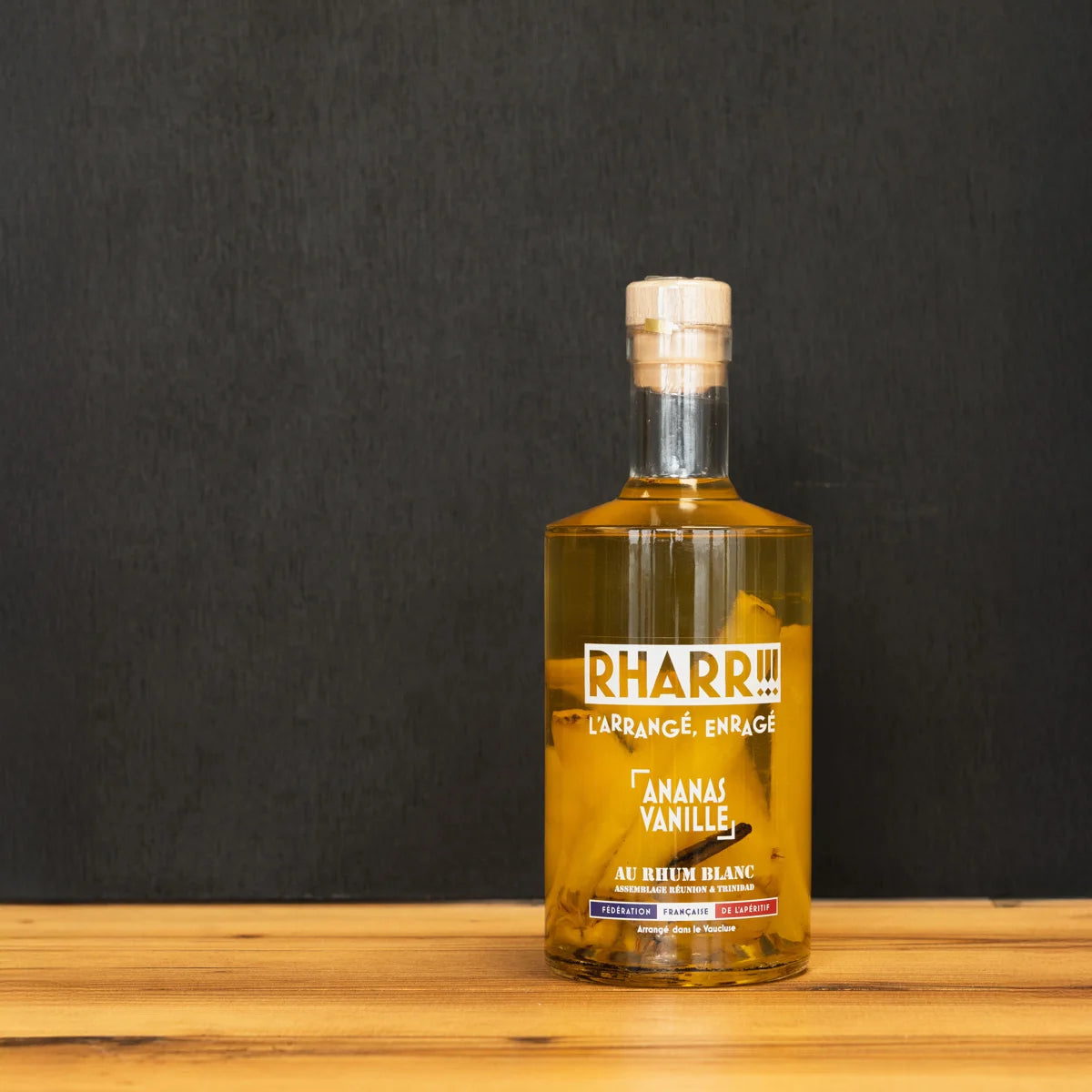 Bouteille de rhum arrangé RHARR !!! Ananas Vanille, macéré artisanalement en France avec morceaux d’ananas et gousses de vanille, posée sur table en bois.