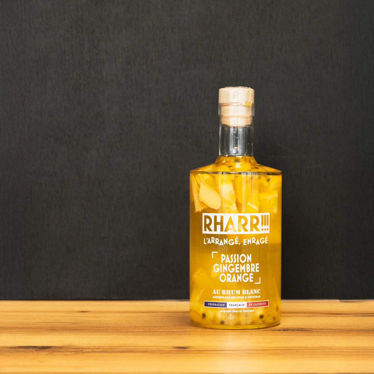 Bouteille de rhum arrangé RHARR !!! Passion Gingembre Orange, macéré artisanalement en France avec morceaux de fruits, posée sur table en bois.