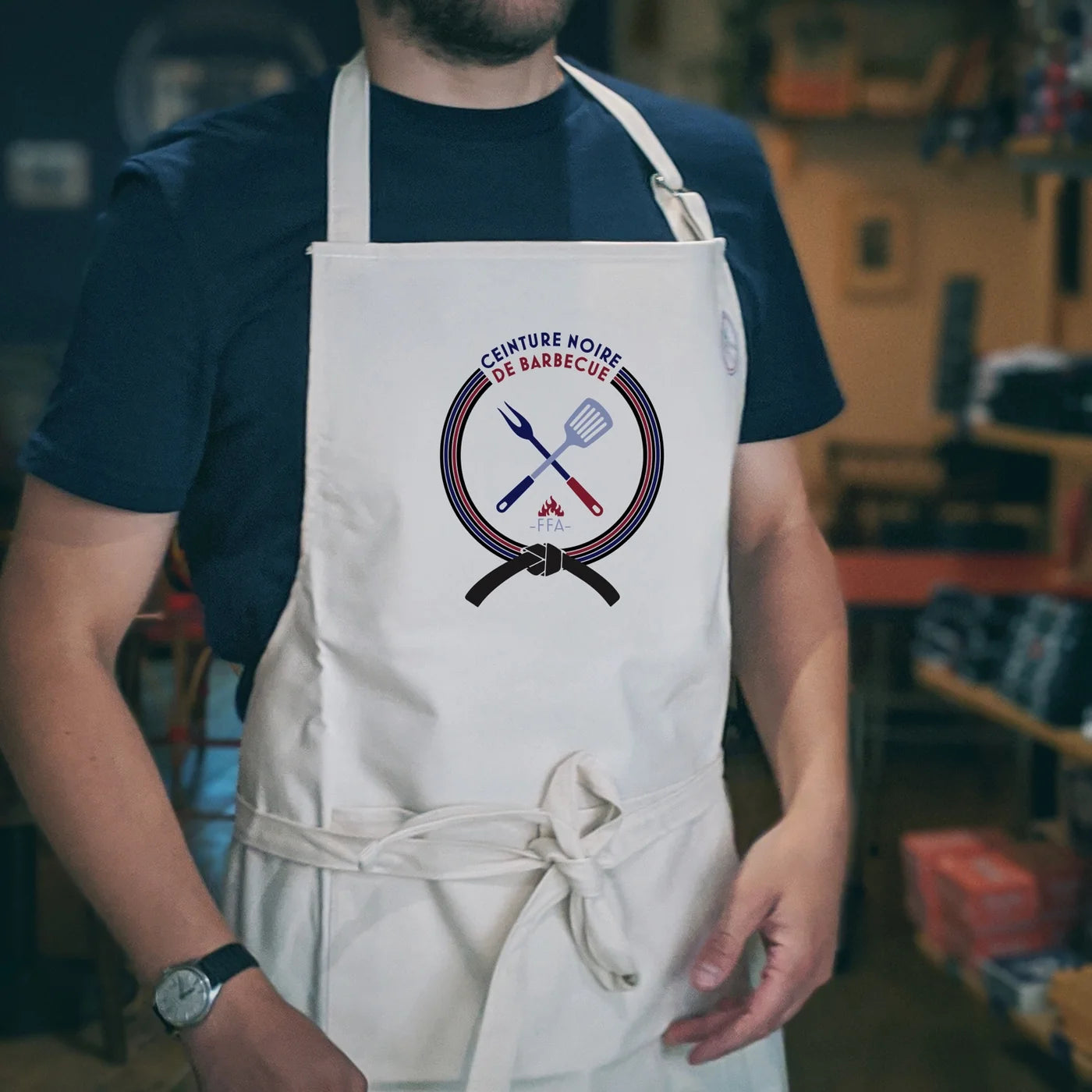 Tablier FFA “Ceinture Noire de Barbecue” porté en intérieur, avec illustration barbecue et ustensiles croisés — ambiance gourmande et conviviale.