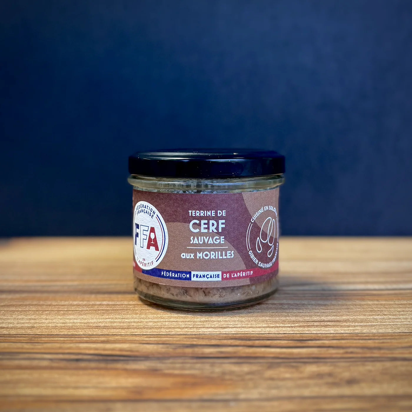 Terrine de cerf sauvage FFA en pot en verre, présentée sur une table en bois, fond bleu foncé, ambiance apéritif naturelle et gourmande.