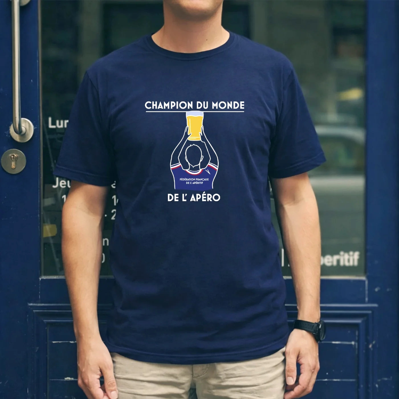T-shirt bleu FFA “Champion du Monde de l’Apéro” porté devant une devanture bleue, avec illustration trophée et verre de bière.