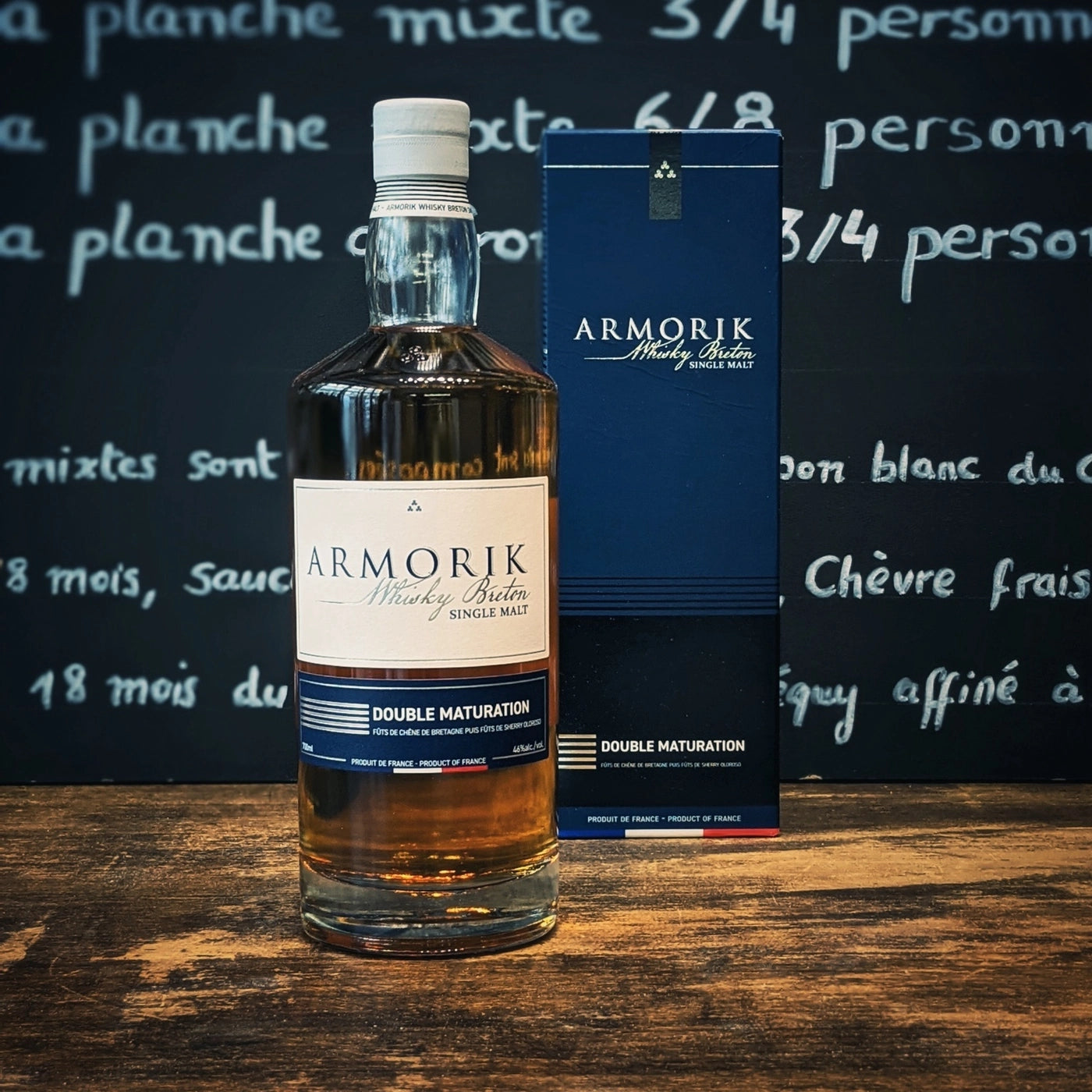 Bouteille de whisky Armorik Double Maturation avec son étui, posée sur une table en bois face à un fond ardoise, ambiance bretonne élégante et chaleureuse.